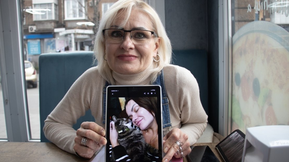 Eine Frau mittleren Alters zeigt frontal ein Bild von einer jungen Frau mit einer Katze im Arm auf einem Tablet. 