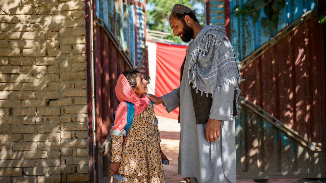 Marwa und ihr Vater stehe vor einem Haus auf einer sonnigen Straße, sie lächeln sich an. Der Vater hat schützend die Hand auf der Schulter seiner Tochter.