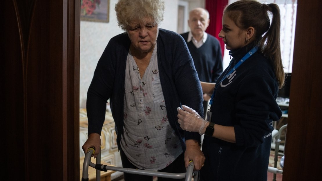 Eine ältere Frau stützt sich auf ihren Rollator. Eine junge Mitarbeiterin von HI stützt sie. 