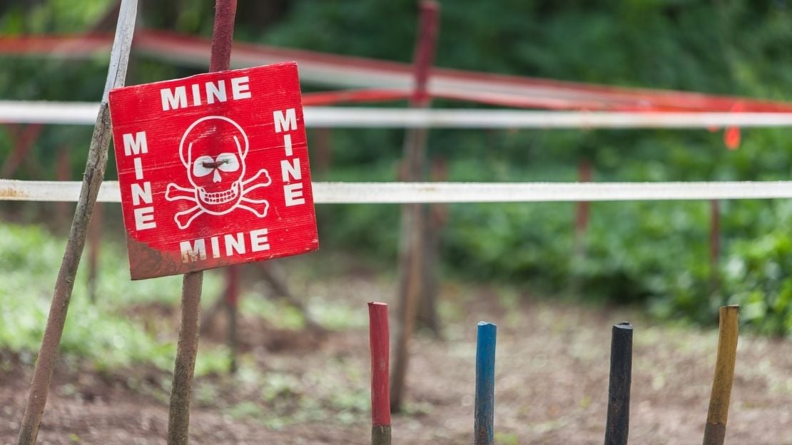 Ein „Danger Mines“-Schild warnt vor der unsichtbaren Gefahr. Daneben stecken Stöcke in der Erde, die den Gefahrenbereich markieren. 