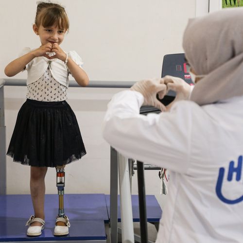 Ein kleines Mädchen mit Beinprothese formt mit den Händen ein Herz und guckt dabei ihre Physiotherapeutin an, die mit ihr Laufen übt.