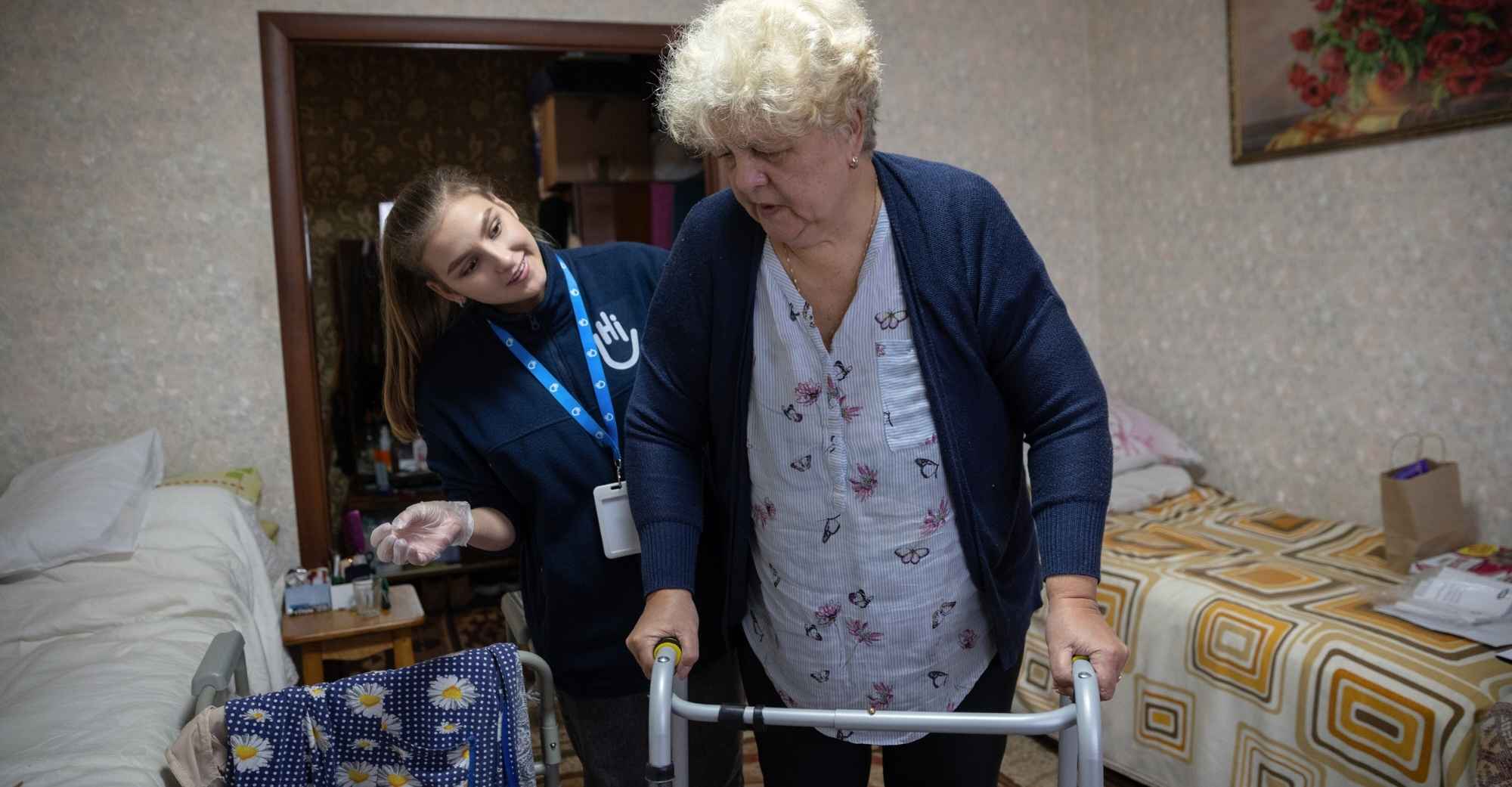 Eine ältere Frau mit kurzen blonden Haaren stützt sich auf ihren Rollator. Hinter ihr steht eine Helferin von Handicap International und lächelt sie an. Im Hintergrund ist ein Wohnzimmer.