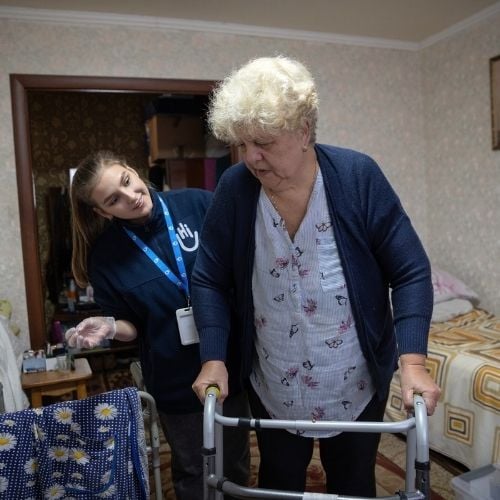 Eine ältere Frau mit kurzen blonden Haaren stützt sich auf ihren Rollator. Hinter ihr steht eine Helferin von Handicap International und lächelt sie an. Im Hintergrund ist ein Wohnzimmer.