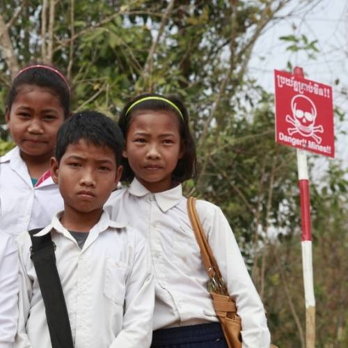 Eine kleine Gruppe Schulkinder steht links und schaut ernst. Rechts neben ihnen steht ein Schild, das mit Totenkopf-Symbol vor Minen warnt.