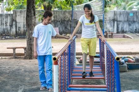 Eine junge Frau mit Beinprothese geht über eine Brücke aus bunten Streben. Neben ihr steht ein Mitarbeiter von Handicap International und hilft ihr.