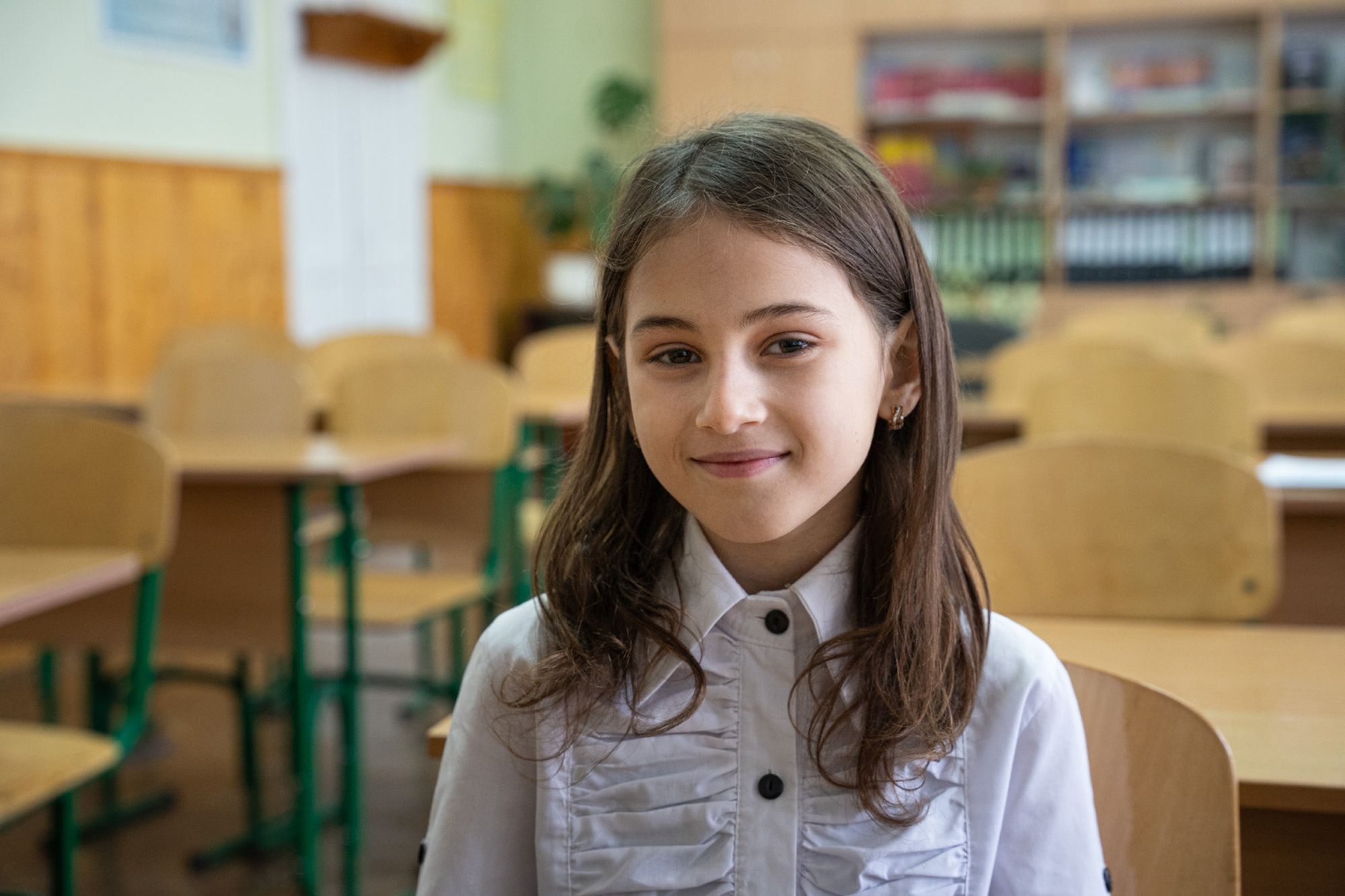 Ein junges Mädchen mit langen braunen Haaren sitzt in einem Klassenraum und lächelt in die Kamera.