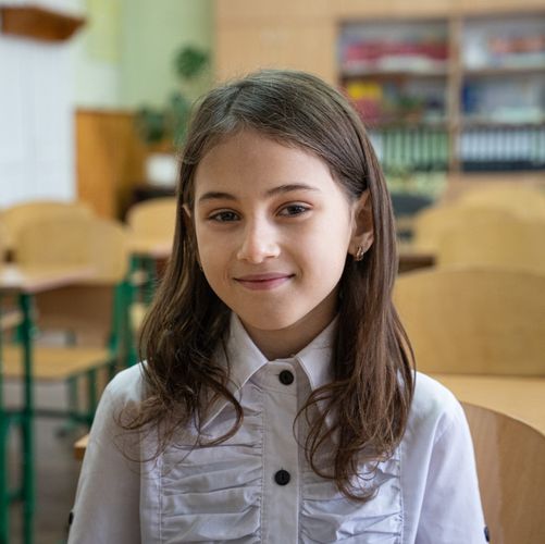 Ein junges Mädchen mit langen braunen Haaren sitzt in einem Klassenraum und lächelt in die Kamera.